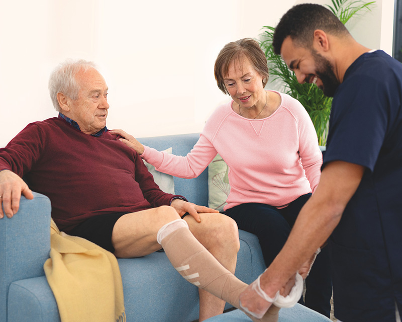 Man seated on a sofa with his leg bandaged while a healthcare worker assists and a women looks on. Man seated on a sofa with his leg bandaged while a healthcare worker assists and a women looks on.